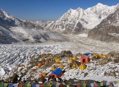 Colorful tents and prayer flags at snowy base camp during Kanchenjunga trekking, with glacier and rugged peaks under a clear blue sky